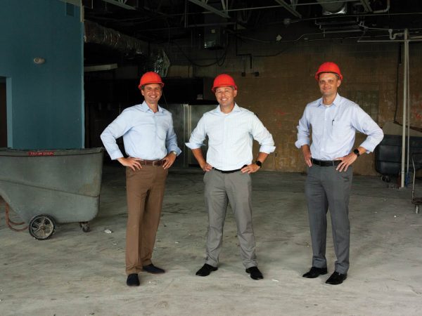 surrounded by construction materials and equipment.|A group of fourteen people stands outdoors in front of a brick building with arched windows. They are dressed in business casual attire