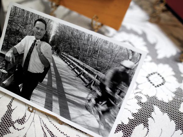 book-filled room. Shes looking at a large photo album on her lap. More albums are on a table in the foreground. The setting is warm and inviting.|A person wearing a floral top and blue pants holds a photo album open to a newspaper clipping with the headline
