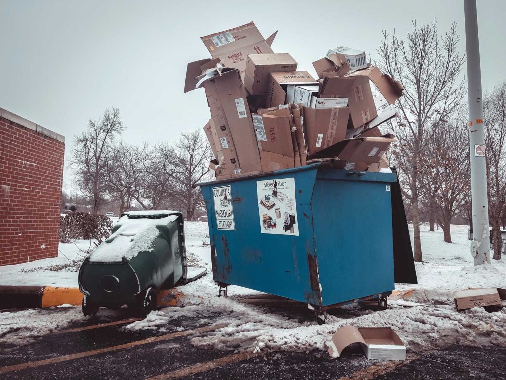 surrounded by a light layer of snow. Leafless trees and a brick building are in the background.|Overflowing dumpsters with cardboard boxes and plastic bags in a snowy parking lot
