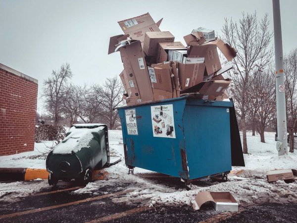 surrounded by a light layer of snow. Leafless trees and a brick building are in the background.|Overflowing dumpsters with cardboard boxes and plastic bags in a snowy parking lot