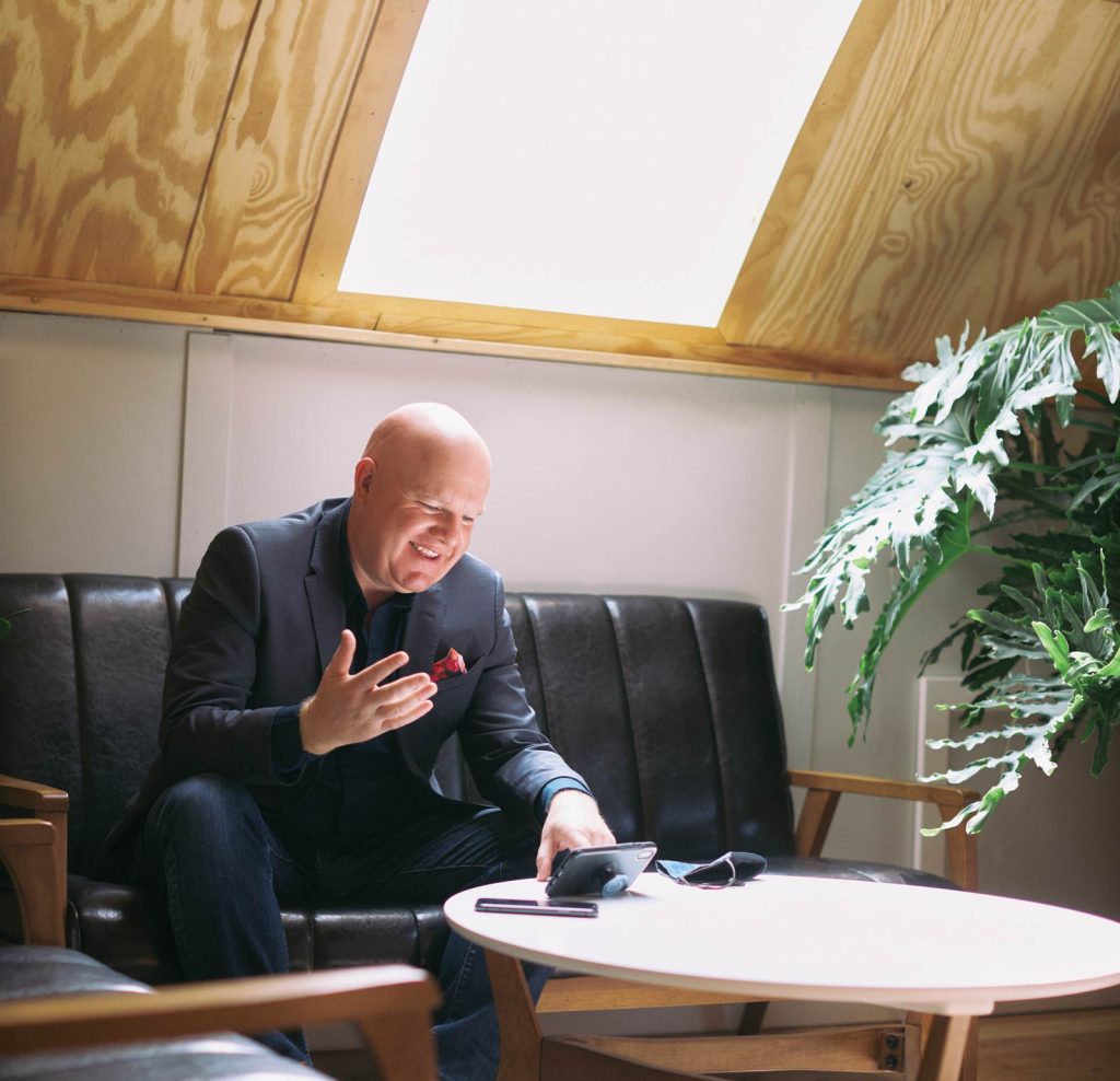 gesturing with his hand in a warmly lit room. He is sitting on a black sofa next to a wooden table