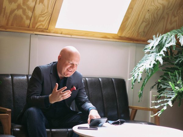 gesturing with his hand in a warmly lit room. He is sitting on a black sofa next to a wooden table