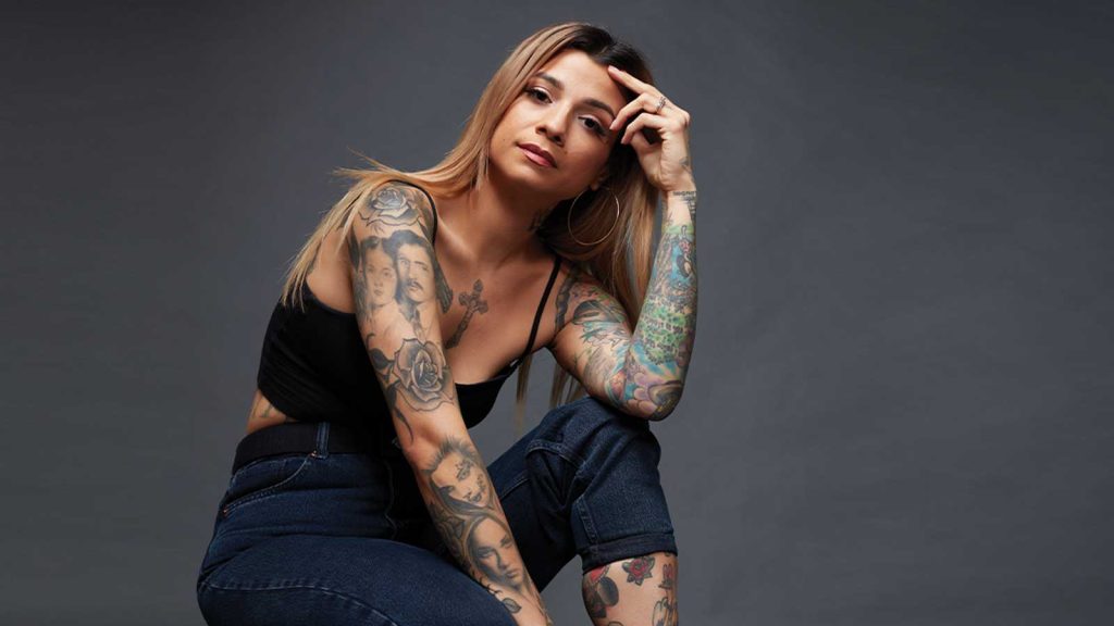 set against a plain dark background.|A collage of three images showing tattoos. The left image displays colorful arm tattoos including a womans face and bird. The center shows a back tattoo of a tiger outline. The right image highlights another colorful arm with various designs.|A person with long hair and hoop earrings gazes into the camera. Their arm is adorned with colorful tattoos