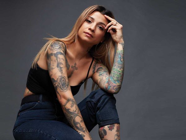 set against a plain dark background.|A collage of three images showing tattoos. The left image displays colorful arm tattoos including a womans face and bird. The center shows a back tattoo of a tiger outline. The right image highlights another colorful arm with various designs.|A person with long hair and hoop earrings gazes into the camera. Their arm is adorned with colorful tattoos