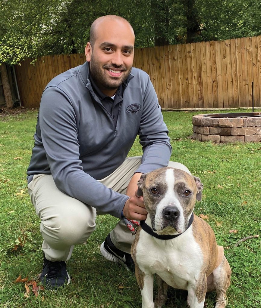 smiling at the camera.|A man is crouching next to a brown and white dog in a grassy backyard with a wooden fence and trees. The man is smiling and wearing a gray jacket and beige pants. The dog is sitting calmly beside him.