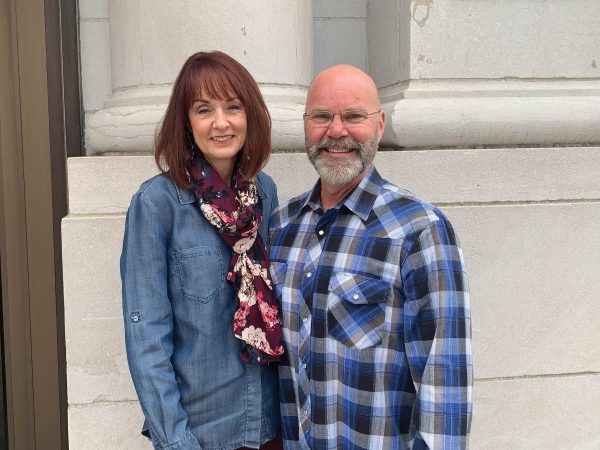 wears a denim jacket and floral scarf. The man has a bald head
