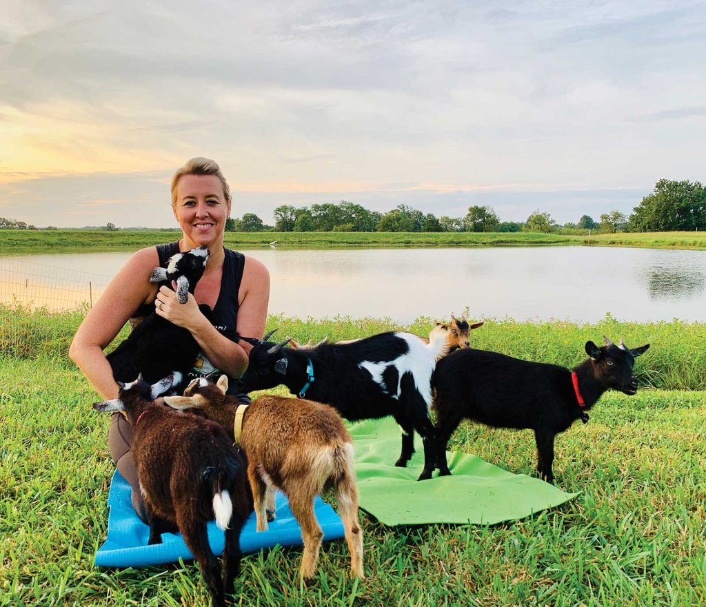 a women on a green yoga mat has a brown goat on her back|A group of people in a outdoor pasture on colorful yoga mats with two goats on their back|in a outdoor pasture