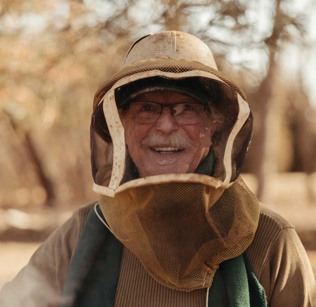 blurred outdoor setting with trees.|A beekeeper in protective gear examines a honeycomb frame in an outdoor setting. Surrounding him are wooden beehives