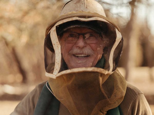 blurred outdoor setting with trees.|A beekeeper in protective gear examines a honeycomb frame in an outdoor setting. Surrounding him are wooden beehives