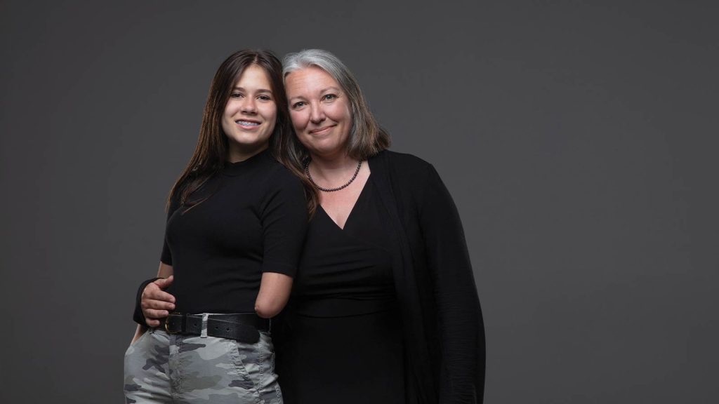 also smiling. They both wear black tops against a gray background.|Two women stand against a gray background