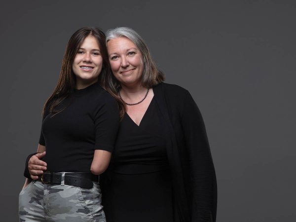 also smiling. They both wear black tops against a gray background.|Two women stand against a gray background