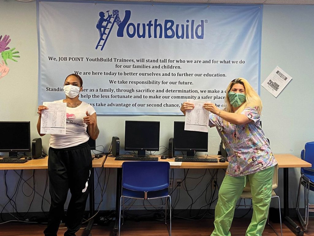 holding printed certificates and smiling. A banner above reads YouthBuild. The setting appears to be a classroom or computer lab.|A person in blue gloves holds an open white foam container with a poppy seed hot dog bun. The hot dog inside is topped with mustard