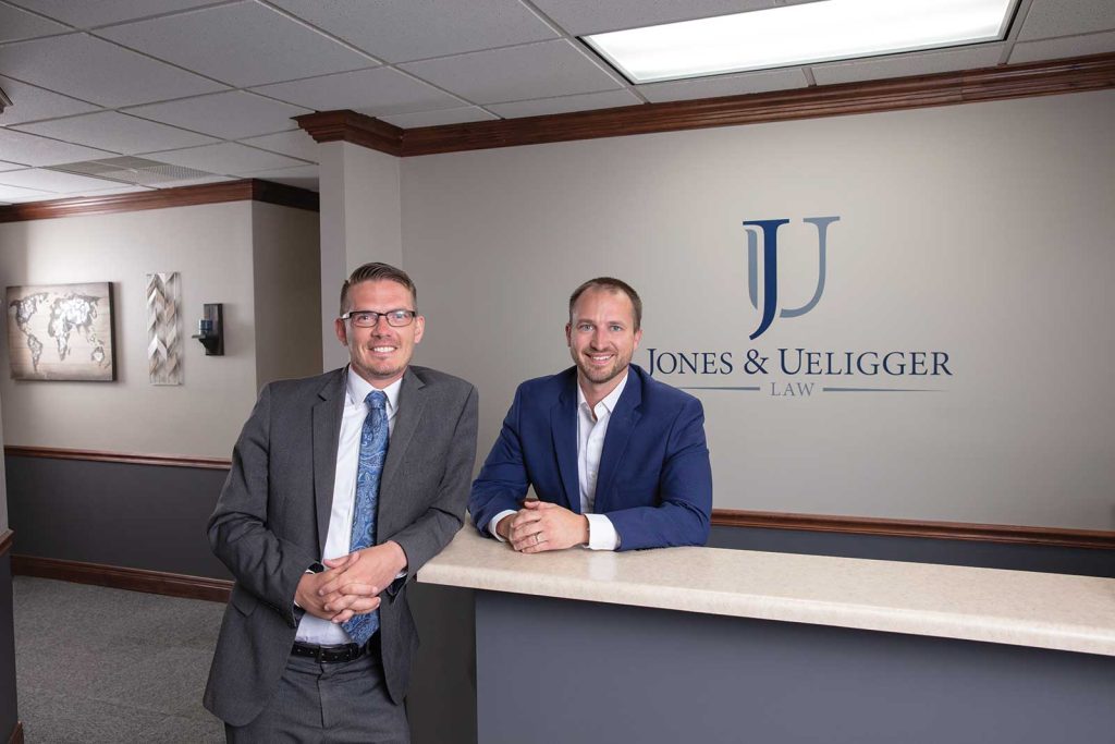 smiling and engaged in conversation. The wall behind them features the company name "Jones & Ueigger Law."|Two men named Jones and Ueligger in business suits smile for a photo in front of a wooden wall. One is wearing a blue suit