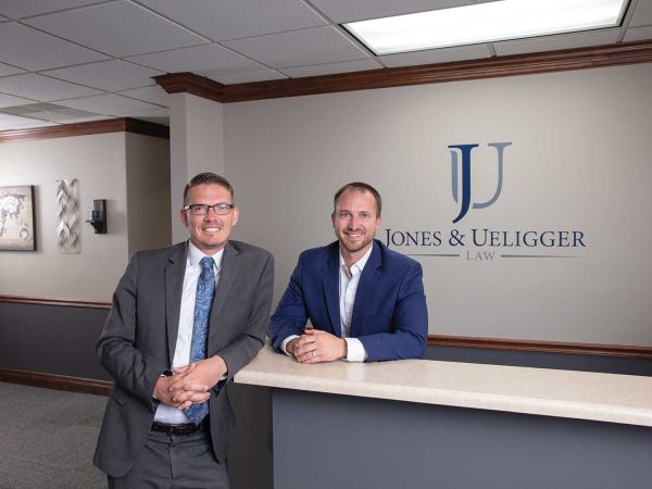 smiling and engaged in conversation. The wall behind them features the company name "Jones & Ueigger Law."|Two men named Jones and Ueligger in business suits smile for a photo in front of a wooden wall. One is wearing a blue suit