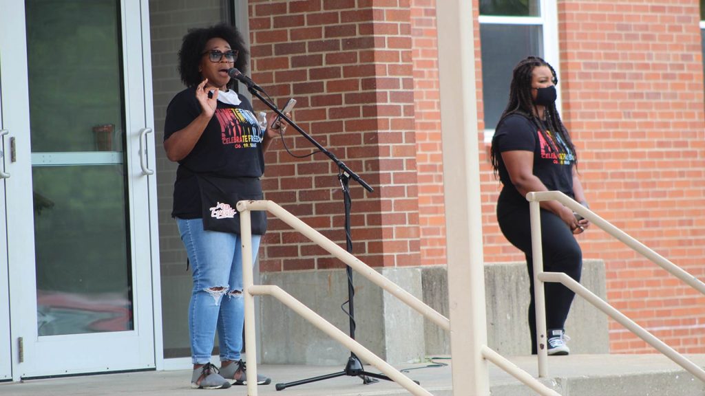 while the other stands nearby. Both wear black T-shirts with colorful text and jeans. The woman speaking has curly hair and the other has long braids and wears a mask.|Two people sit in folding chairs by a parked vehicle