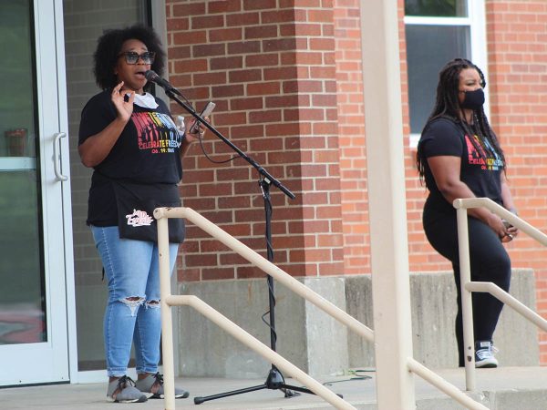 while the other stands nearby. Both wear black T-shirts with colorful text and jeans. The woman speaking has curly hair and the other has long braids and wears a mask.|Two people sit in folding chairs by a parked vehicle