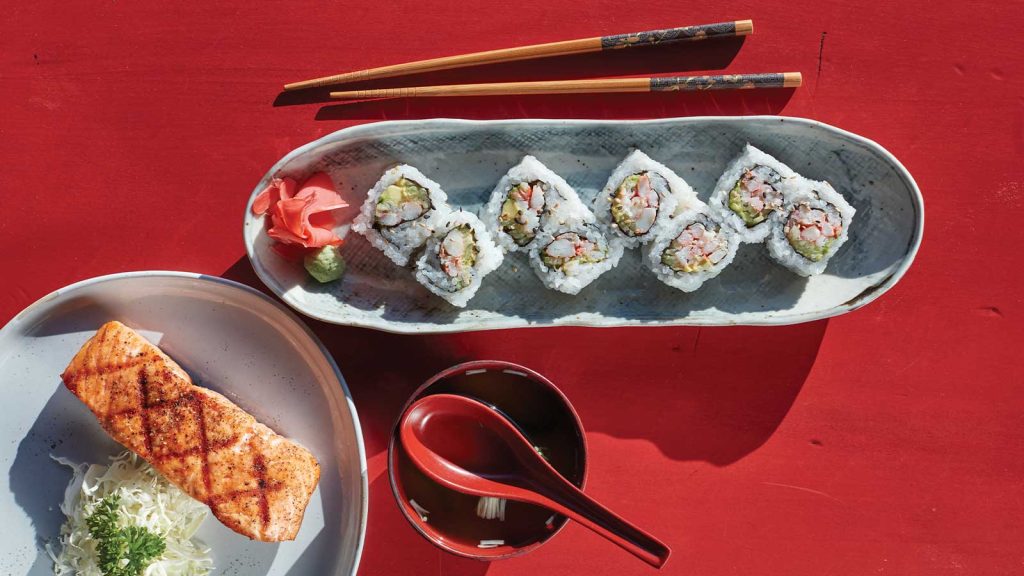 all on a red surface with a pair of chopsticks beside them.|Left image: Sushi rolls on a ceramic dish with pickled ginger and wasabi