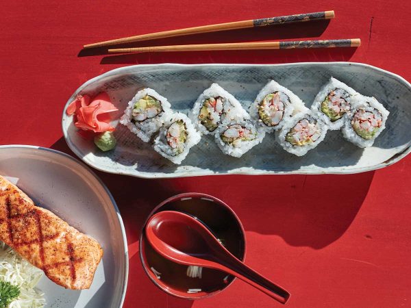 all on a red surface with a pair of chopsticks beside them.|Left image: Sushi rolls on a ceramic dish with pickled ginger and wasabi
