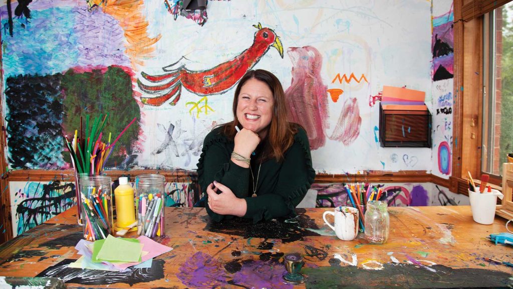 leaning on a colorful arts and crafts table. The room is filled with vibrant art on the walls