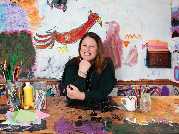 leaning on a colorful arts and crafts table. The room is filled with vibrant art on the walls