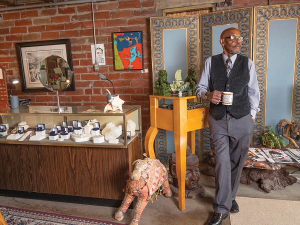 holding a mug and leaning against a small table with decorations. Nearby are a display case with jewelry