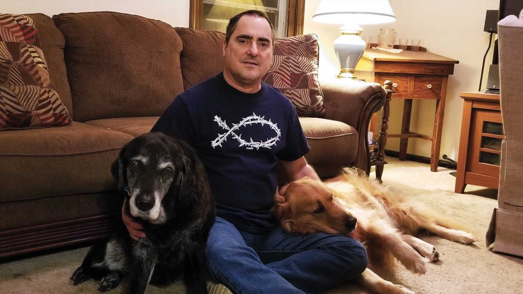 wearing a navy blue t-shirt and jeans. He is surrounded by two dogs: a black dog on his left and a golden retriever on his right. The sofa