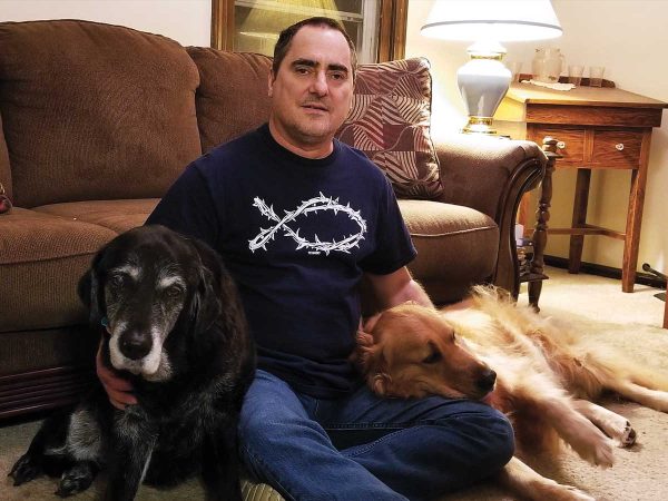 wearing a navy blue t-shirt and jeans. He is surrounded by two dogs: a black dog on his left and a golden retriever on his right. The sofa
