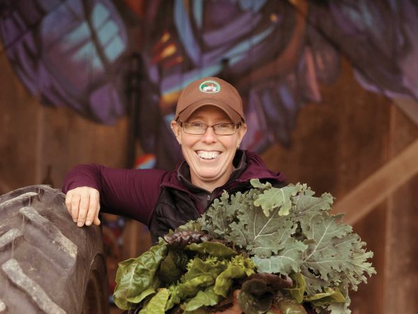 smiling and holding leafy greens. There is a colorful mural in the background.|A bundle of colorful Swiss chard with vibrant green leaves and multicolored stems in shades of red