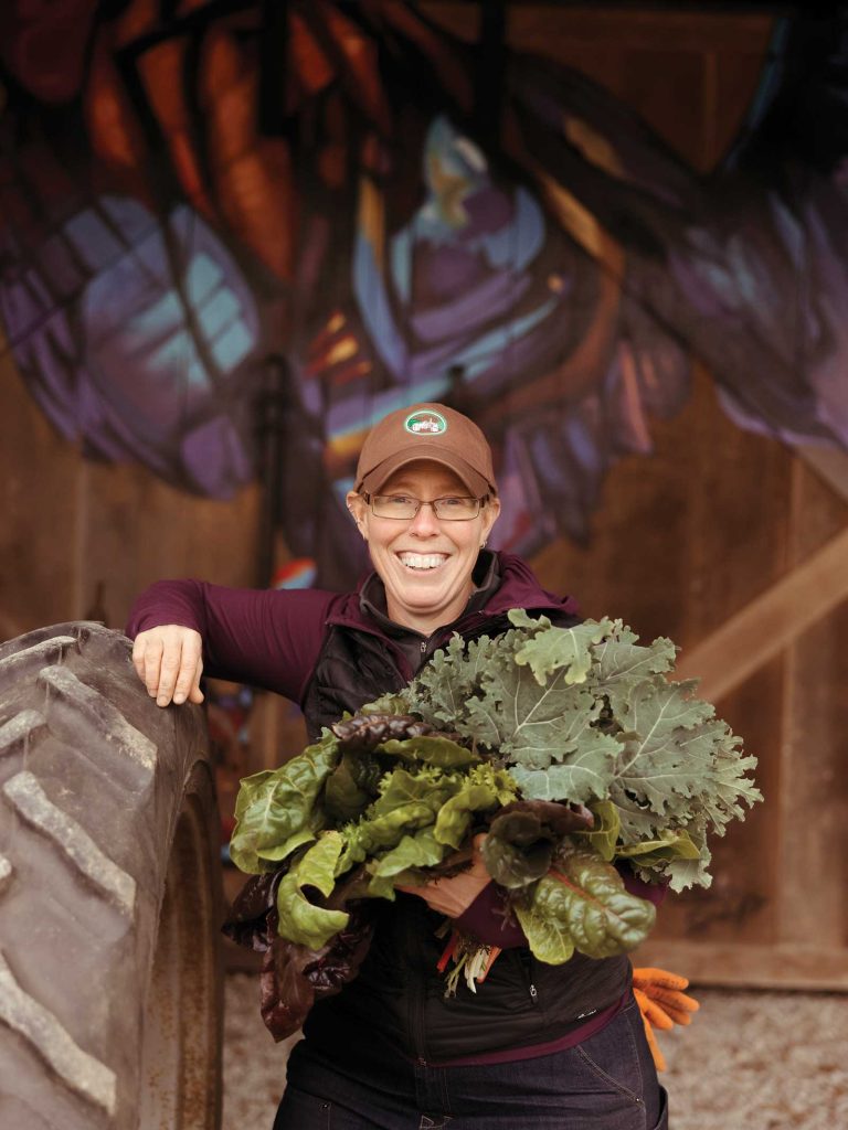 smiling and holding leafy greens. There is a colorful mural in the background.|A bundle of colorful Swiss chard with vibrant green leaves and multicolored stems in shades of red