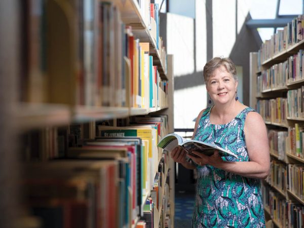 holding an open book. She is wearing a sleeveless dress with a blue and green pattern and smiling at the camera. Shelves filled with books surround her