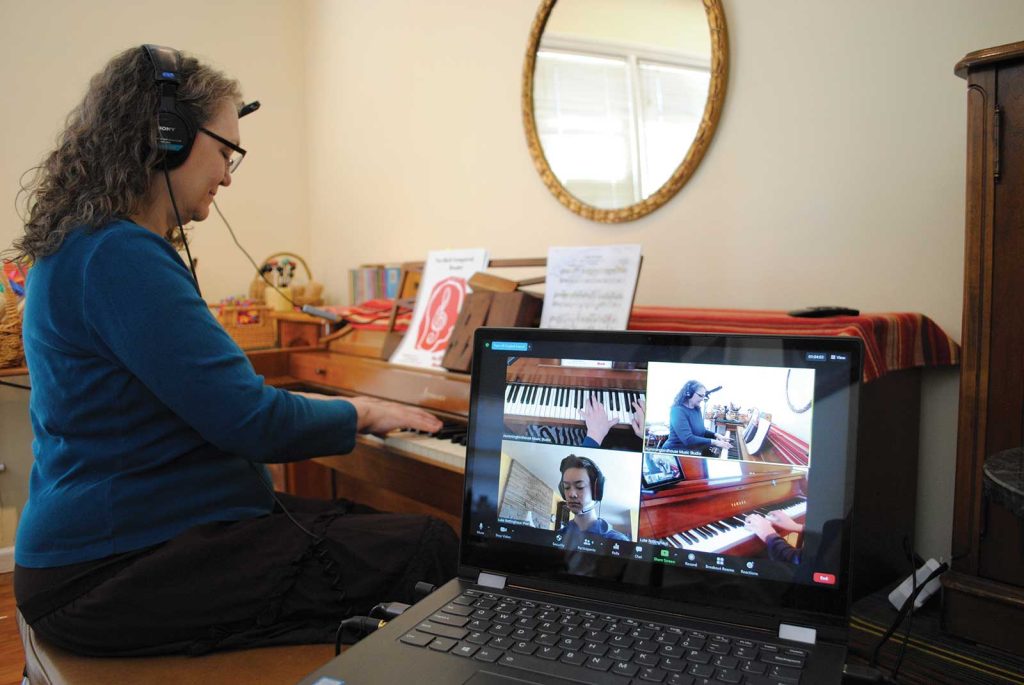 engaged in an online video call on a laptop. The screen displays three people participating in a virtual music session. A mirror and sheet music are visible in the background.|A woman with long curly hair is sitting on a stool