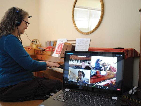 engaged in an online video call on a laptop. The screen displays three people participating in a virtual music session. A mirror and sheet music are visible in the background.|A woman with long curly hair is sitting on a stool