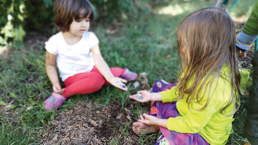 while the child on the right wears a yellow top and a colorful skirt. A partial view of another person is in the background.|Left: A young girl with a leafy crown stands outdoors