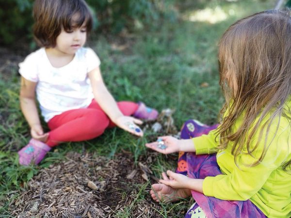 while the child on the right wears a yellow top and a colorful skirt. A partial view of another person is in the background.|Left: A young girl with a leafy crown stands outdoors