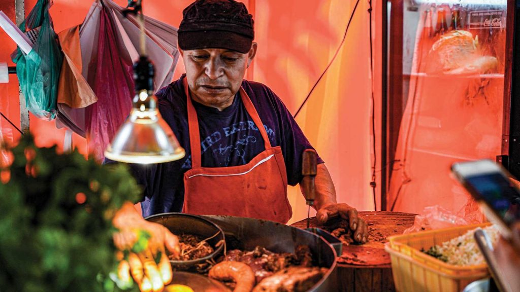 with green herbs visible in the foreground. The background is warmly lit in orange tones.|A street vendor stands beside a colorful cart filled with fresh fruit and snacks