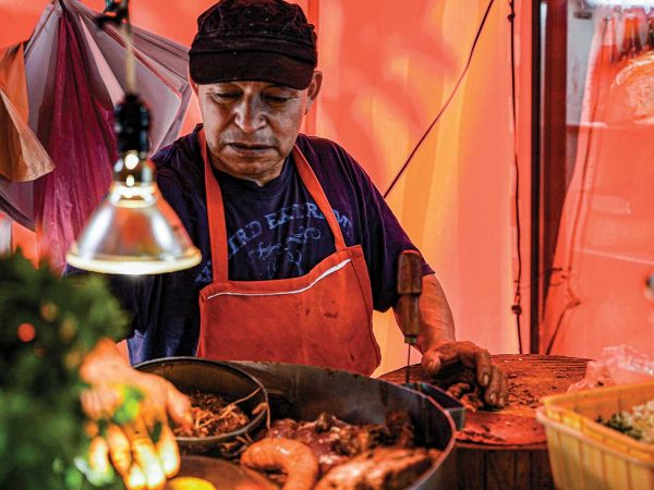 with green herbs visible in the foreground. The background is warmly lit in orange tones.|A street vendor stands beside a colorful cart filled with fresh fruit and snacks