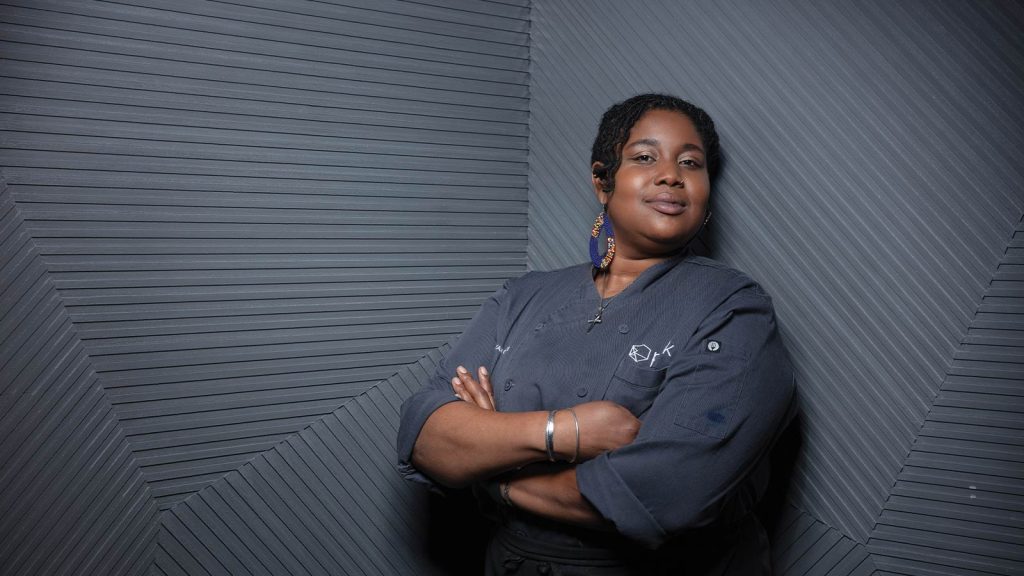 and a plated dish is visible on the counter. She is wearing a dark chefs jacket and gloves.|Chef in a commercial kitchen
