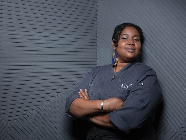 and a plated dish is visible on the counter. She is wearing a dark chefs jacket and gloves.|Chef in a commercial kitchen