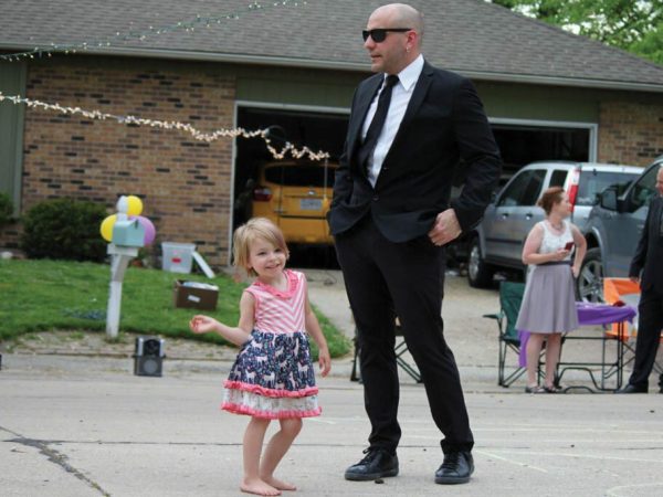 a man in a black suit and sunglasses stands with his hands in his pockets. The background shows a house