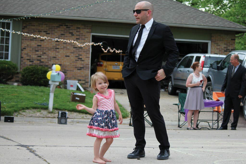 a man in a black suit and sunglasses stands with his hands in his pockets. The background shows a house