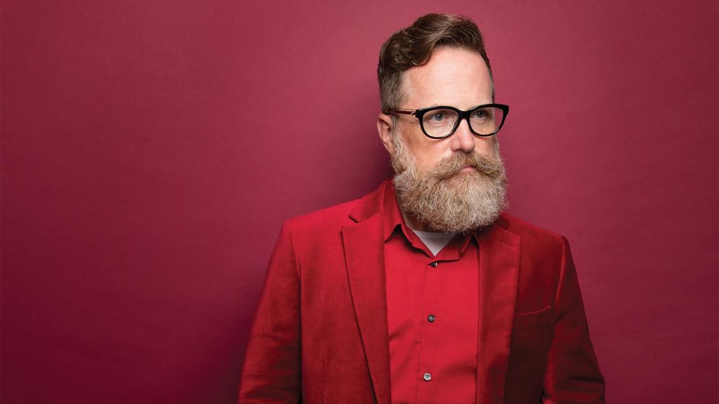 looking to the side.|A person with glasses and a full beard looks directly at the camera. They are wearing a light pink suit jacket and a shirt