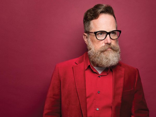 looking to the side.|A person with glasses and a full beard looks directly at the camera. They are wearing a light pink suit jacket and a shirt