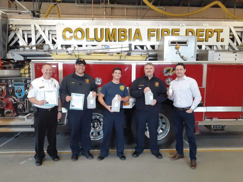 while another man in a uniform and a man in a white shirt and jeans smile next to them. They are inside a fire station.|Three people stand smiling beside a stack of boxes containing healthcare supplies. They are in an office with a University of Missouri System sign in the background. Two people wear masks