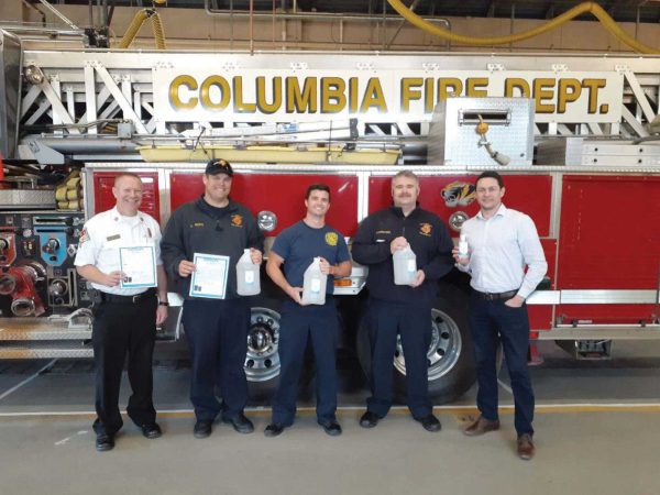 while another man in a uniform and a man in a white shirt and jeans smile next to them. They are inside a fire station.|Three people stand smiling beside a stack of boxes containing healthcare supplies. They are in an office with a University of Missouri System sign in the background. Two people wear masks