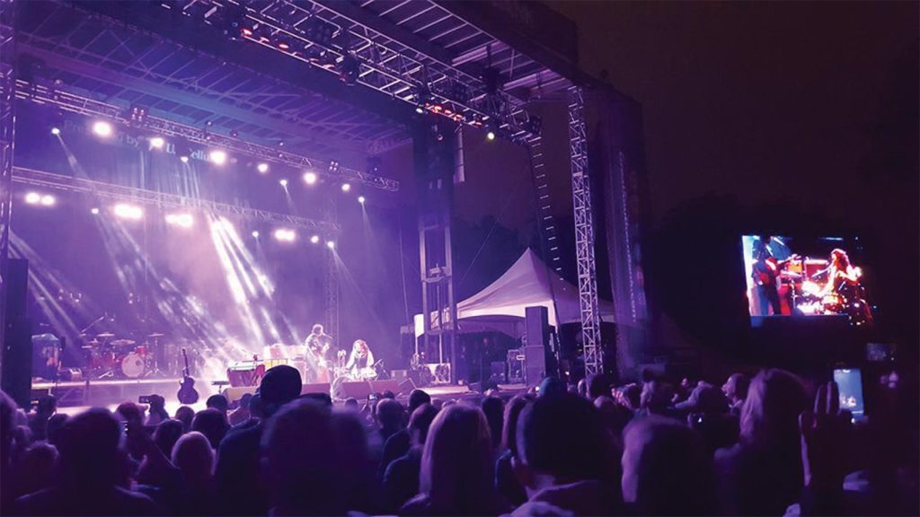 silhouetted in the foreground. Bright spotlights illuminate the performers. A large screen shows the performance to the side.|A scenic view of a wooden deck overlooking a river at sunset. Two people sit on chairs