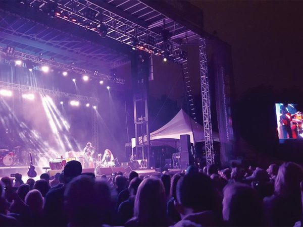 silhouetted in the foreground. Bright spotlights illuminate the performers. A large screen shows the performance to the side.|A scenic view of a wooden deck overlooking a river at sunset. Two people sit on chairs