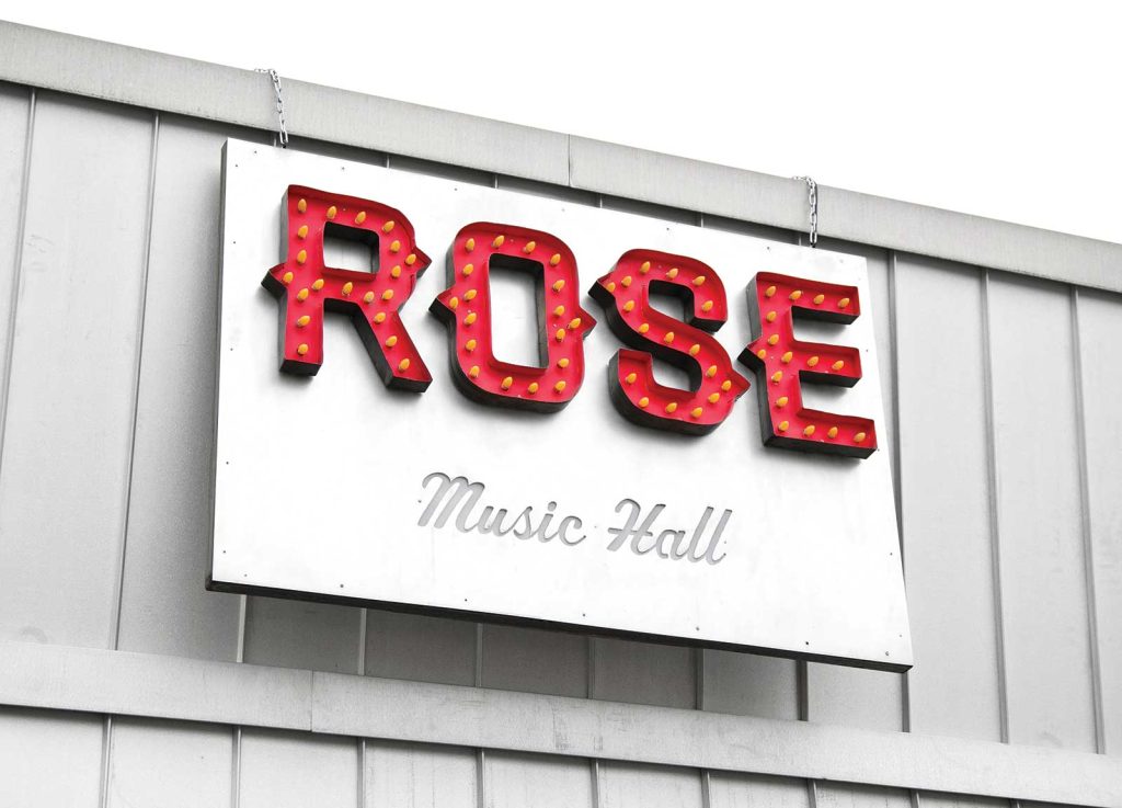 cursive font. The sign is mounted on a light gray wall with vertical panels.|A person in a flannel shirt and cap leans out of a pizza shops open service window. Neon signs above read Pizza and By The Slice. The building is brick