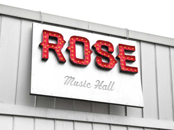 cursive font. The sign is mounted on a light gray wall with vertical panels.|A person in a flannel shirt and cap leans out of a pizza shops open service window. Neon signs above read Pizza and By The Slice. The building is brick