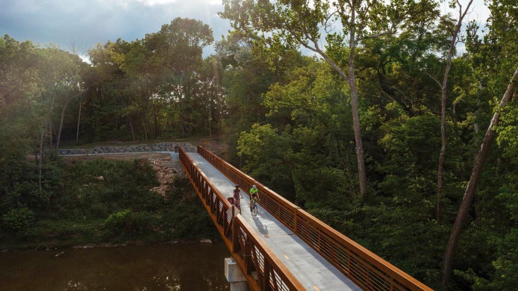 narrow bridge surrounded by lush green trees. The bridge spans over a calm river