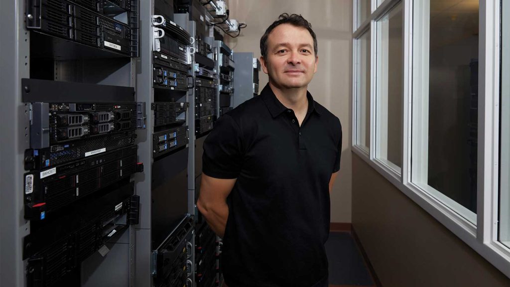 surrounded by racks of computer servers. White-framed windows line the right side of the room. He is smiling and has his hands behind his back.|Smiling woman in a floral blouse stands in a modern office environment with people working at computers in the background. The atmosphere is professional and welcoming.|A person wearing a blue checkered shirt stands smiling in an office environment with cubicles and computers. The background shows other people working at their desks.